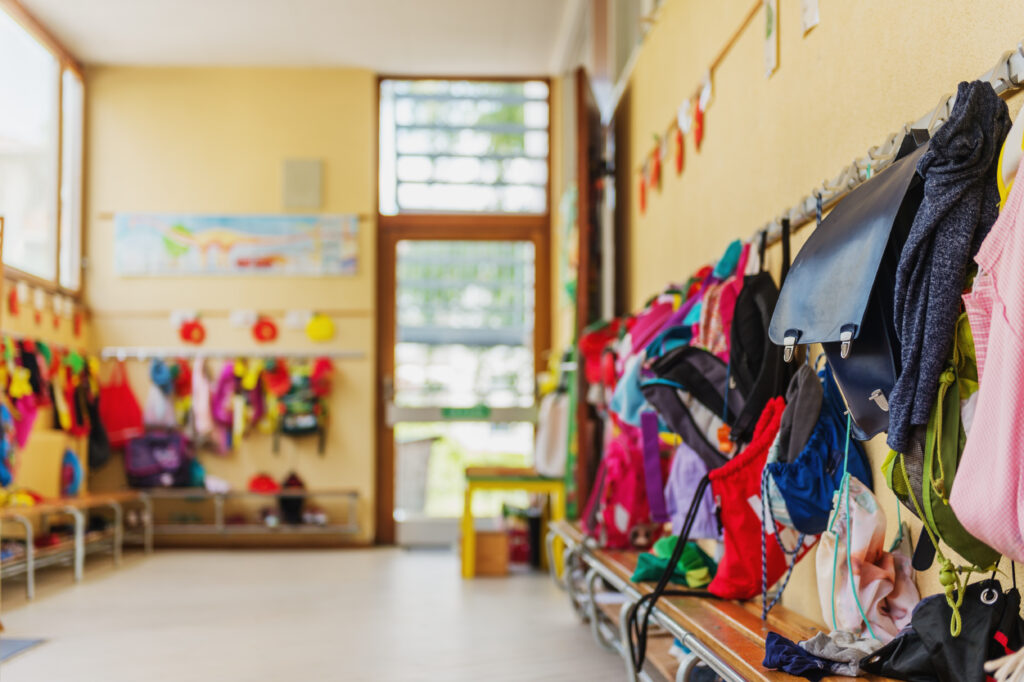 Vestiaire d’établissement scolaire avec manteaux accroché à des porte-manteaux et cartables d’enfants posés sur des bancs.