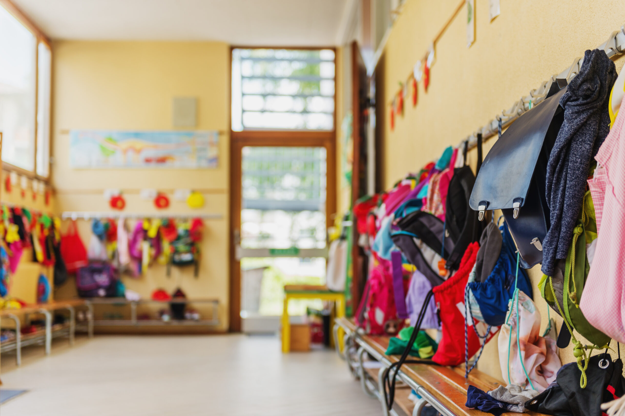 Vestiaire d’établissement scolaire avec manteaux accroché à des porte-manteaux et cartables d’enfants posés sur des bancs.